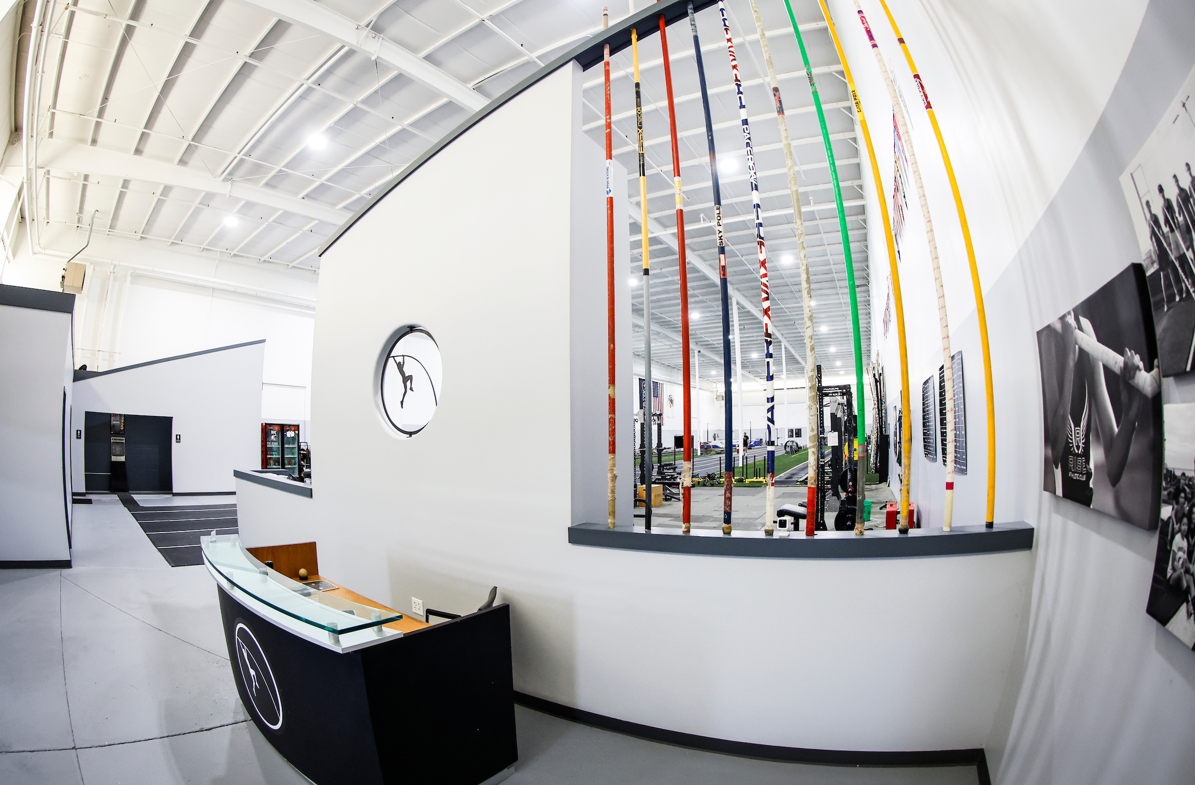 RISE Pole Vault A modern gym interior with a reception desk in the foreground, poles leaning against a railing, and workout equipment visible in the background. The walls are white with a high ceiling, and framed photos are displayed on the wall.
